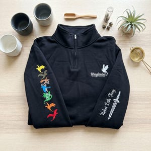 Op de afbeelding: Zwarte trui met een kwart ritssluiting. De linkermouw heeft geborduurde draken in verschillende kleuren. De rechter mouw heeft een witte dolk en de tekst "Violent Little Thing". Op de borst staat een witte draak en het woord "Wingleader".