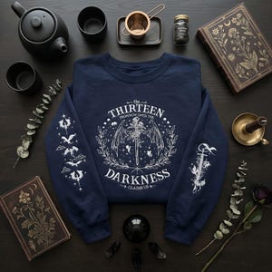 May include: Dark navy sweatshirt featuring white graphic text and illustrations. The design includes the words "the THIRTEEN FROM NOW UNTIL THE DARKNESS CLAIMS US +" in a circular emblem with a sword, wings, and floral elements. Sleeves feature additional white illustrations.