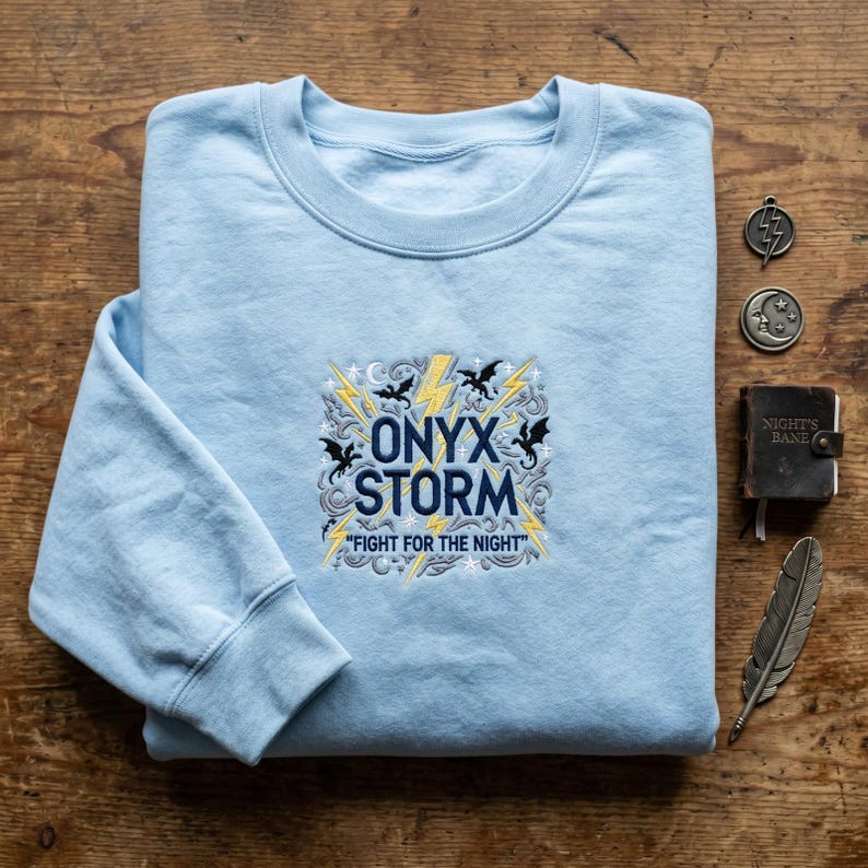 May include: A light blue crewneck sweatshirt featuring an embroidered design with the text ONYX STORM FIGHT FOR THE NIGHT, surrounded by yellow lightning bolts, black dragon silhouettes, and grey swirling patterns on the front center.