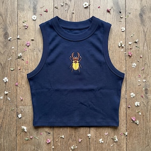 May include: A navy blue ribbed sleeveless crop top featuring a central embroidered graphic of a stag beetle with a brown head and yellow wing covers. The garment has a high crew neckline and wide arm openings, showcasing a textured fabric.