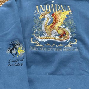 Puede incluir: Una sudadera azul con un drag&oacute;n bordado y texto. El drag&oacute;n es dorado, naranja y blanco, con la palabra "ANDARNA" encima. El texto "I WILL NOT LET THEM BURN YOU" est&aacute; debajo. La manga izquierda tiene un dise&ntilde;o bordado m&aacute;s peque&ntilde;o y el texto "I will not die today."