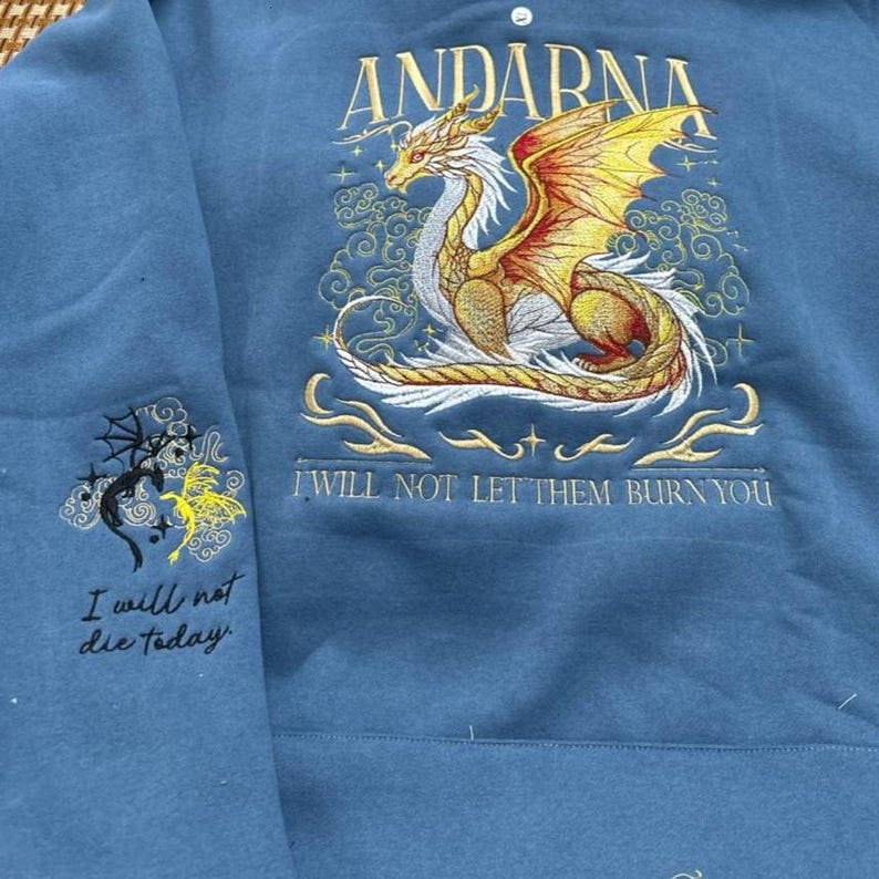 Puede incluir: Una sudadera azul con un drag&oacute;n bordado y texto. El drag&oacute;n es dorado, naranja y blanco, con la palabra "ANDARNA" encima. El texto "I WILL NOT LET THEM BURN YOU" est&aacute; debajo. La manga izquierda tiene un dise&ntilde;o bordado m&aacute;s peque&ntilde;o y el texto "I will not die today."