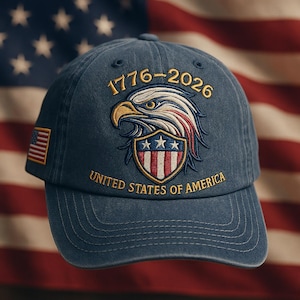 May include: A navy blue baseball cap with embroidered details. The cap features a patriotic design with an eagle, stars, and stripes, along with the text "1776-2026" and "United States of America". An American flag patch is on the side.