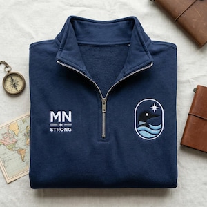 May include: Navy blue quarter-zip sweatshirt with the text "MN STRONG" and a patch featuring a loon and star. The sweatshirt has a silver zipper and a stand-up collar. A compass, map, and leather journal are in the background.
