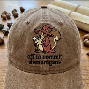 May include: Brown baseball cap with an embroidered squirrel wearing a wizard hat and robe, reading a book. The text "off to commit shenanigans" is below the squirrel. The cap is made of a soft, worn material.
