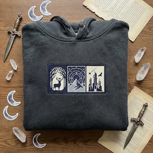 May include: Dark gray hoodie with a three-panel embroidered design. The panels depict a deer in a forest, a mountain range with a starry sky, and a cityscape with a bird. The hoodie has a drawstring hood.