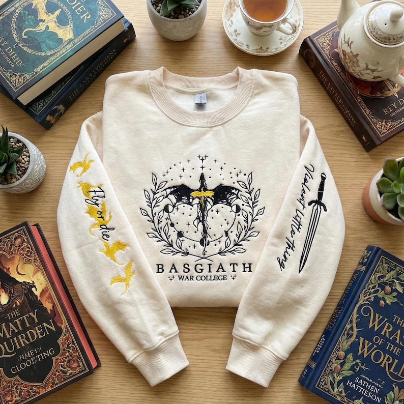May include: Cream-colored sweatshirt with embroidered design. Features a central emblem with a sword and wings, the text "BASGIATH WAR COLLEGE", and the phrases "Fly or die" and "Violent little thing" on the sleeves. Surrounded by books and plants.