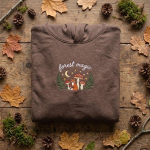 May include: A brown hooded sweatshirt featuring an embroidered design of a fox, mushrooms, ferns, a crescent moon, and stars. The text "forest magic" is stitched in white above the central graphic, which is centered on the chest area.