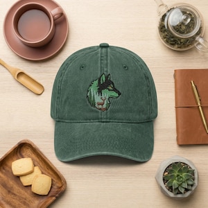 May include: A green baseball cap with an embroidered design of a wolf's head, trees, and a deer. The cap is made of a soft, worn material and has a curved brim. The design features black, green, and brown colors.