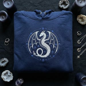 Peut inclure: Un sweat à capuche bleu marine avec un motif de dragon blanc brodé. Le dragon est entouré d'un cadre décoratif avec des étoiles. Ce sweat-shirt décontracté est un vêtement confortable.