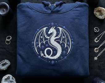 May include: A navy blue hooded sweatshirt featuring a detailed white dragon design within a circular frame. The dragon has outstretched wings and is surrounded by stars. The sweatshirt is a comfortable, casual garment.
