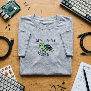 May include: A heather gray t-shirt with the text "CTRL + SHELL" and a cartoon turtle pressing a CTRL key. The shirt is surrounded by computer components, keyboards, and cables on a wooden surface.