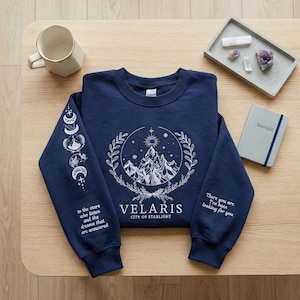 Peut inclure: Sweat-shirt bleu marine avec un motif blanc de montagne et de ciel. Il comporte le texte "VELARIS CITY OF STARLIGHT" et les phrases "to the stars who listen and the dreams that are answered" et "There you are. I've been looking for you."