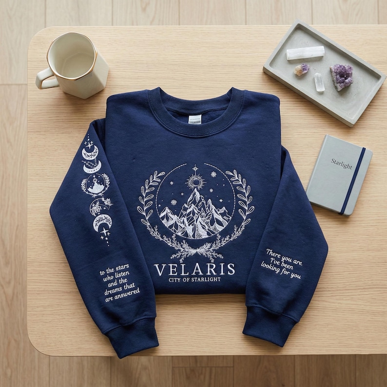 Peut inclure: Sweat-shirt bleu marine avec un motif blanc de montagne et de ciel. Il comporte le texte "VELARIS CITY OF STARLIGHT" et les phrases "to the stars who listen and the dreams that are answered" et "There you are. I've been looking for you."