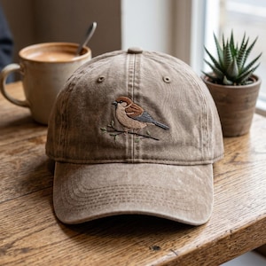Peut inclure: Une casquette de baseball beige à la finition vieillie, ornée d'une broderie représentant un moineau brun et blanc perché sur une fine branche. La casquette présente une calotte structurée, une visière courbée et des œillets cousus.