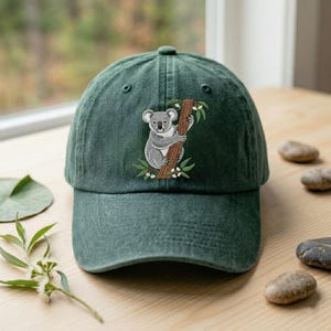 Könnte beinhalten: Dunkelgrüne Baseballkappe mit einer Stickerei eines Koalas, der sich an einem Ast festhält. Der Koala ist grau und weiß, der Ast braun. Die Kappe besteht aus einem weichen, strukturierten Stoff.