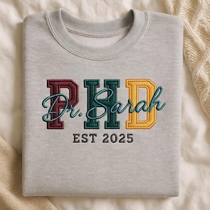 May include: A pale grey sweatshirt featuring embroidered text. The word "PhD" is in maroon, green, and gold block letters. The name "Dr. Sarah" is in teal script, and "EST 2025" is below in brown.