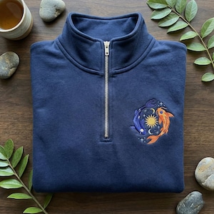 May include: Navy blue quarter-zip sweatshirt with an embroidered design of two koi fish, one blue and one orange, encircling a sun and moon. The sweatshirt has a high collar and a silver zipper. The design is surrounded by stars.