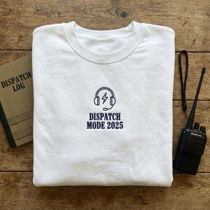 May include: A white crew neck sweatshirt with the words "DISPATCH MODE 2025" embroidered in navy blue, along with a headset and lightning bolt graphic. A black walkie-talkie and a "DISPATCH LOG" notebook are also visible.