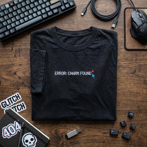 May include: A black short-sleeved t-shirt featuring white embroidered text that reads ERROR: CHARM FOUND, accompanied by a small red dot and a blue pixelated computer mouse cursor icon positioned at the end of the text on the chest area.