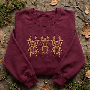 May include: A maroon sweatshirt featuring a gold embroidered design of three stag beetles. The beetles are centered on the front of the crew neck sweatshirt. The design is a detailed outline of the insect.