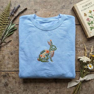 Peut inclure: Un sweat-shirt bleu clair à col rond orné d'une broderie centrale représentant la silhouette d'un lapin composée de diverses feuilles vertes, de tiges et de petites fleurs orange et jaunes. Le vêtement possède un col côtelé.