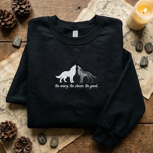 May include: Black sweatshirt with embroidered images of two wolves, one white and one gray, facing each other. The text "Be wary. Be clever. Be good." is embroidered below the wolves. The sweatshirt is displayed on a wooden surface with a map and candle.