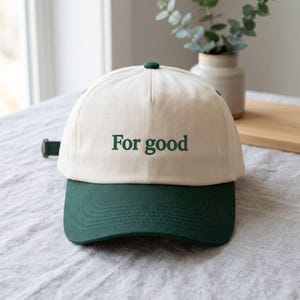 May include: A two-tone baseball cap featuring a cream-coloured crown and a dark green brim. The front of the crown displays the text For good in dark green lettering. The cap includes a dark green adjustable strap with a metal buckle on the side.