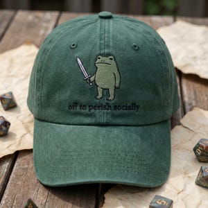 Könnte beinhalten: Grüne Baseballkappe mit einer gestickten Frosch-Illustration, der ein Schwert hält. Unter dem Frosch ist der Text "off to perish socially" gestickt. Die Kappe besteht aus einem weichen Stoff und hat einen gebogenen Schirm.