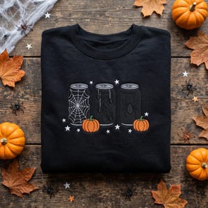 May include: A black crewneck sweatshirt featuring an embroidered graphic of three beverage cans. The cans are decorated with a spiderweb, small spiders, and a large spider. Two orange pumpkins and several white stars surround the cans.