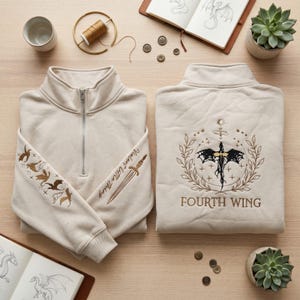 May include: Beige quarter-zip sweatshirt with embroidered dragon and sword designs. The sleeve features dragons and the text "Violent Little Thing." The back displays a sword and dragon emblem with "FOURTH WING" below.