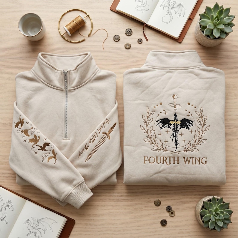 May include: Beige quarter-zip sweatshirt with embroidered dragon and sword designs. The sleeve features dragons and the text "Violent Little Thing." The back displays a sword and dragon emblem with "FOURTH WING" below.