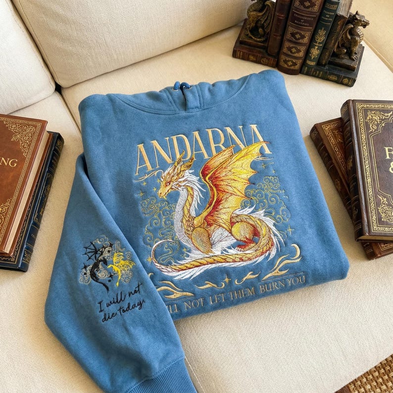 Puede incluir: Una sudadera con capucha azul con la palabra "ANDARNA" bordada en oro. Un dise&ntilde;o detallado de drag&oacute;n con alas doradas y naranjas aparece en la parte delantera. La manga tiene un dise&ntilde;o de drag&oacute;n m&aacute;s peque&ntilde;o y el texto "I will not die today".