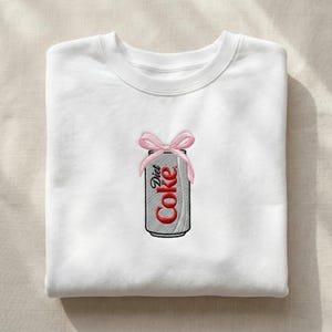May include: White sweatshirt with an embroidered Diet Coke can design. The can is silver with red text and a pink bow at the top. The sweatshirt is folded neatly on a neutral surface.