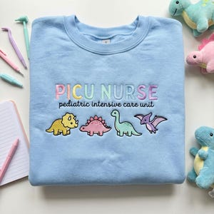 May include: A light blue sweatshirt embroidered with the words "PICU NURSE" in pastel colors, with the words "pediatric intensive care unit" below. Four cartoon dinosaurs are embroidered below the text.