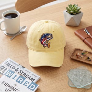 May include: A pale yellow baseball cap featuring an embroidered graphic of a fish with a pattern of the American flag, including blue stars on the head and red and white stripes on the body, centred on the front panel of the six-panel crown.