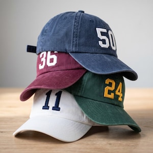 May include: Four baseball caps stacked on top of each other. The caps are navy blue, burgundy, green, and white. Each cap has a different number embroidered on the front in a contrasting color: 50, 36, 24, and 11.