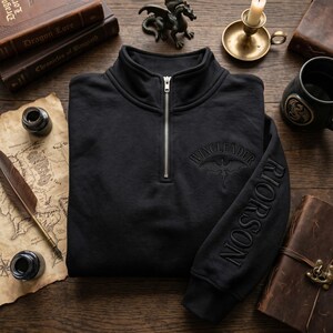May include: Black quarter-zip sweatshirt with the word "WINGLEADER" and a dragon graphic embroidered on the chest. The sleeve features the name "RIORSON" embroidered in a vertical line. The sweatshirt has a silver zipper.