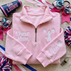 Könnte beinhalten: Ein hellrosa Sweatshirt mit Viertelreißverschluss und den weiß gestickten Worten "Cheer Mom". Eine Cheerleader-Grafik und der Name "Ava" sind ebenfalls gestickt. Cheerleading-Zubehör, darunter eine Megaphon, Schleifen und Pompons, umgeben das Sweatshirt.
