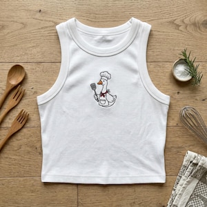 May include: A white ribbed tank top featuring a central embroidered graphic of a goose wearing a chef hat, a red neckerchief, and an apron while holding a spatula. The design is rendered in black, white, orange, and red thread on the fabric.