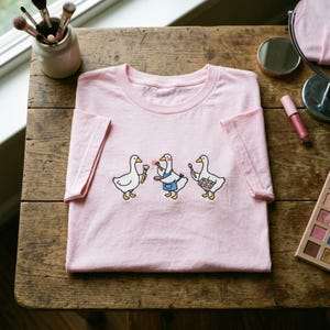 May include: A pale pink t-shirt with a playful illustration of three geese, each holding makeup brushes and palettes. The geese are wearing aprons and holding makeup tools. The shirt is displayed on a wooden surface.