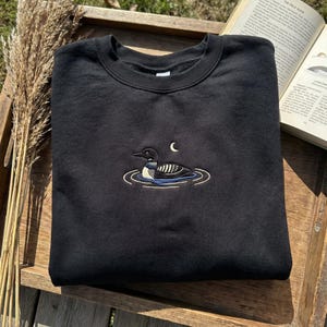 May include: Black crew neck sweatshirt featuring an embroidered design of a loon swimming in water under a crescent moon. The loon has black, white, and blue details. The sweatshirt is folded on a wooden tray.