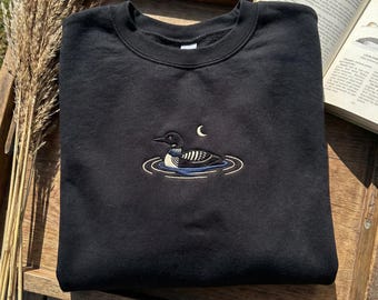 May include: Black crewneck sweatshirt with an embroidered design of a loon swimming in water under a crescent moon. The loon has black, white, and blue details. The sweatshirt is folded neatly on a wooden tray.