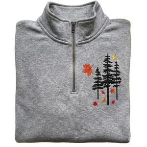 May include: A heather gray quarter-zip pullover featuring an embroidered design of three black pine trees surrounded by small, colorful autumn leaves in shades of orange, red, and yellow. The garment has a high collar and a metal zipper closure.