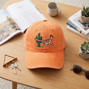 May include: An orange baseball cap featuring an embroidered graphic of a roadrunner bird running past a green saguaro cactus. The cap has a structured crown, a curved brim, and a button on top, showcasing a desert-themed design on the front panel.