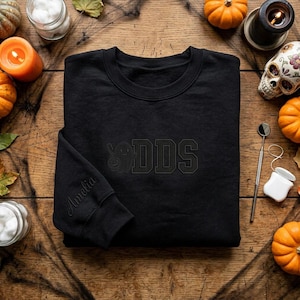 May include: A black crewneck sweatshirt featuring tonal black embroidery on the chest with a ghost graphic and the letters DDS. The name Amelia is embroidered in a cursive script on the sleeve cuff. The garment has a ribbed collar and cuffs.