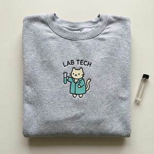 May include: A heather gray sweatshirt with a crew neck. The sweatshirt features a cartoon cat wearing a lab coat and holding a test tube, with the words "LAB TECH" above the cat. A small glass vial is to the right.