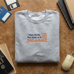 May include: A grey sweatshirt featuring the embroidered phrase "I Keep All My Dad Jokes In A DAD-A-BASE" in blue and orange, with a floppy disk icon. The garment is presented on a wooden surface, accompanied by computer accessories.
