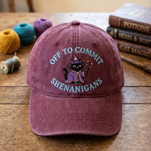 May include: A maroon baseball cap with the embroidered phrase "OFF TO COMMIT SHENANIGANS." The cap features a black cat wearing a wizard hat and robe, holding a wand. The background includes spools of thread and books.