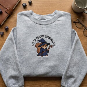 May include: A heather gray sweatshirt featuring an embroidered design of a squirrel dressed as a wizard, holding a staff. The text "OFF TO COMMIT SHENANIGANS" is arched above the squirrel. The sweatshirt is made of a soft material.